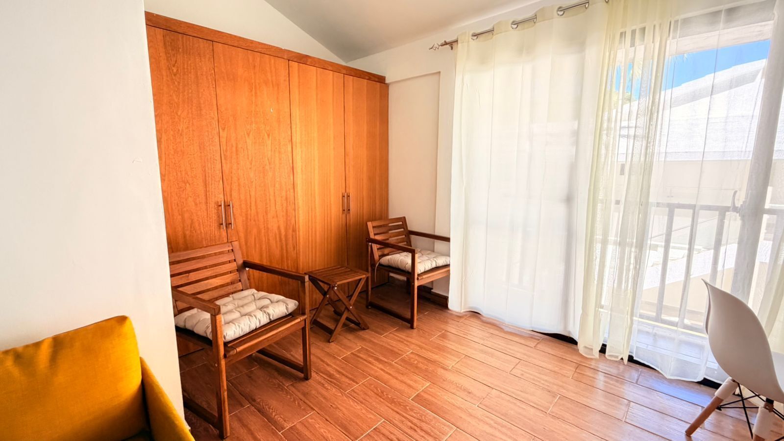 A room with wooden flooring, a wardrobe, two wooden chairs, a small table, and a balcony with a white curtain.