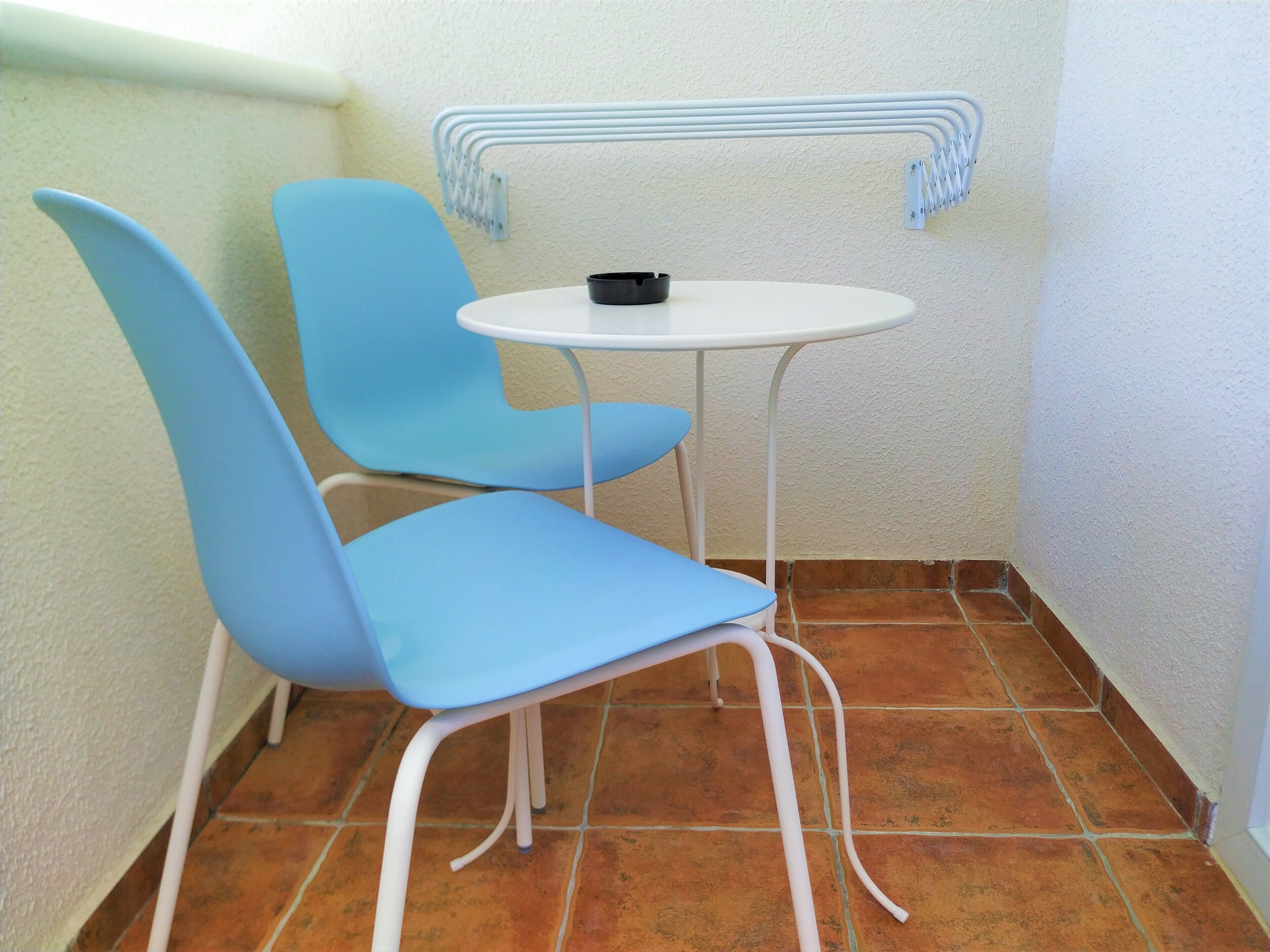 A small, enclosed room with two blue chairs and a white table, featuring a tiled floor and a white wall with a metal rack.