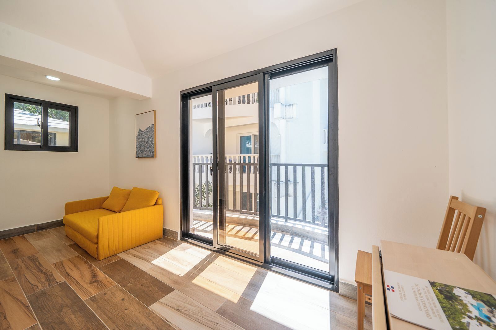 Una habitación con un sofá amarillo, una mesa de madera y un ventanal corredero que da a un balcón.