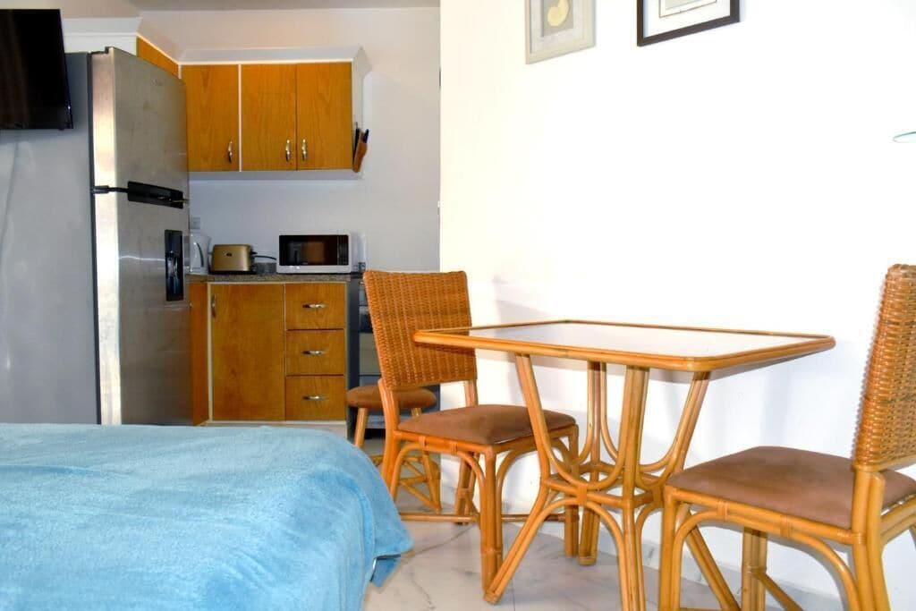 A compact room with a bed, a dining table with chairs, a kitchenette with wooden cabinets, and a microwave.