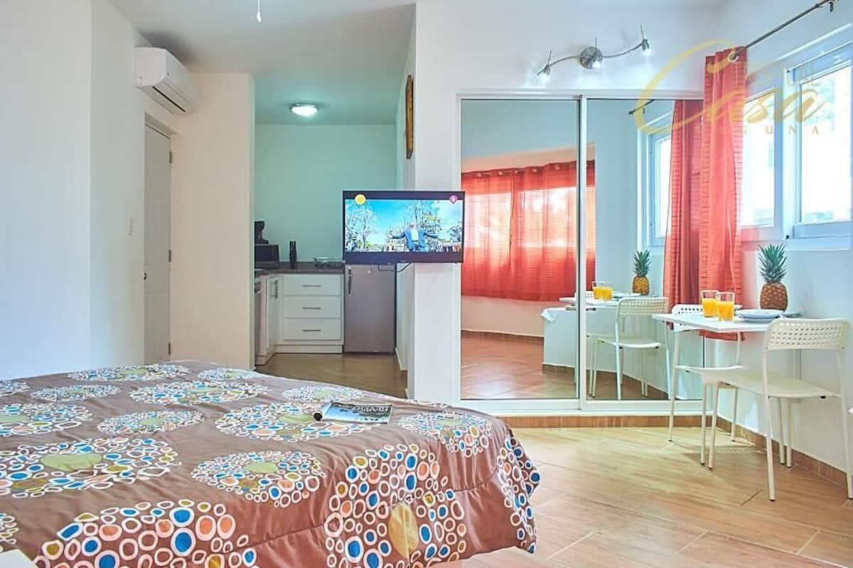 A bedroom with a bed, a TV, a kitchenette, and a dining area with chairs and a table.