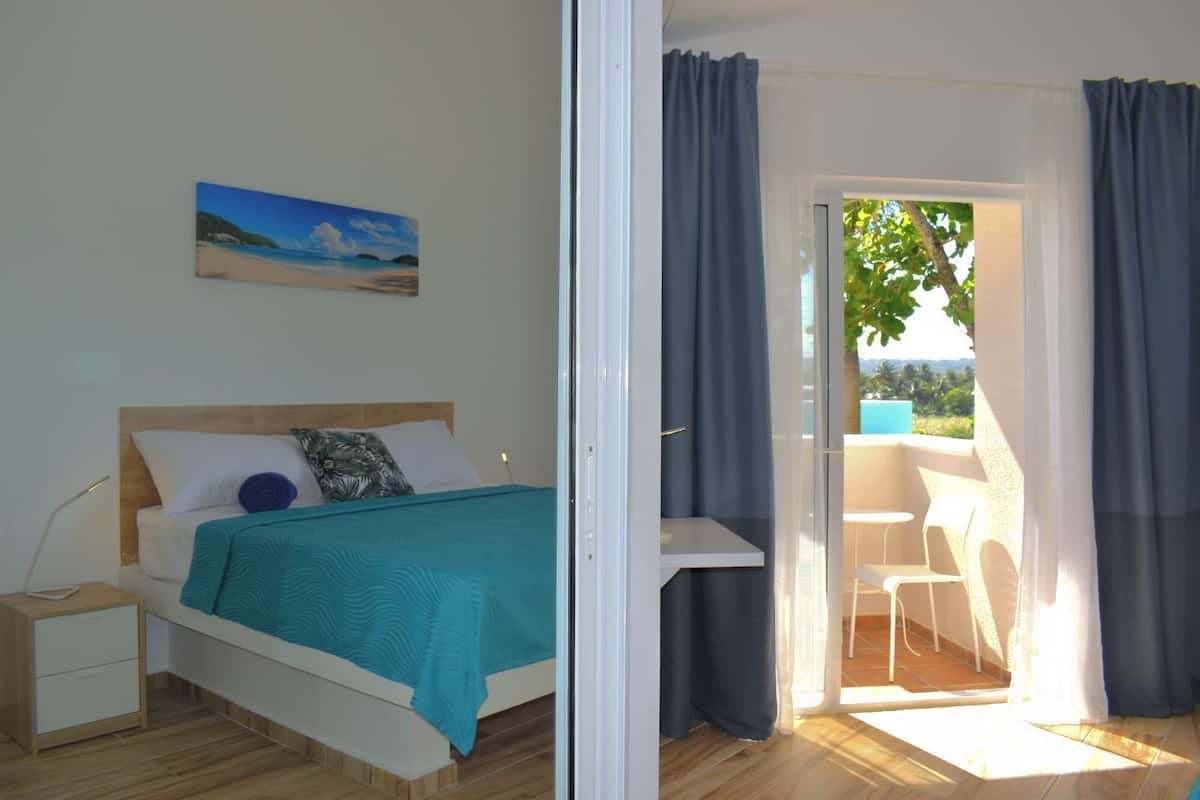 A bedroom with a bed, a nightstand, a painting, and a view of an outdoor area with a table and chairs.