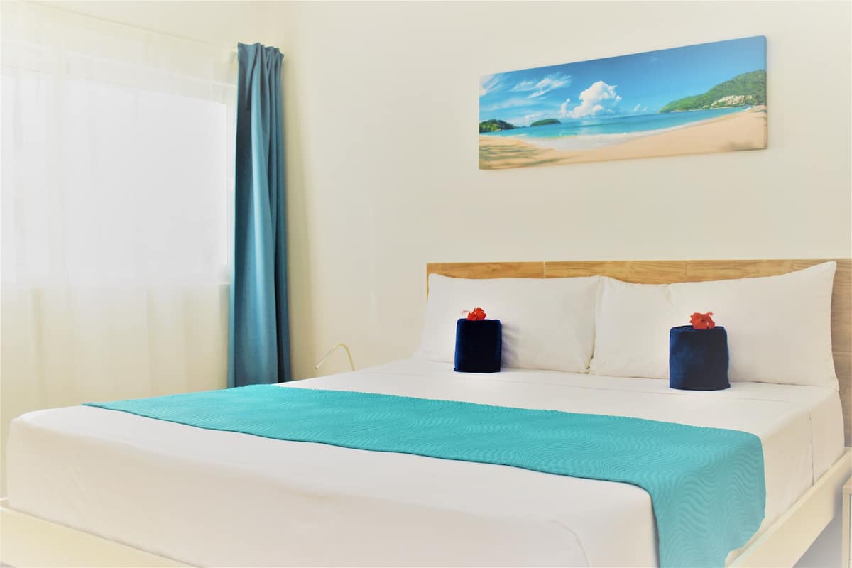 A neatly made bed with a teal bedspread and two pillows, a painting of a beach scene on the wall, and a window with curtains.