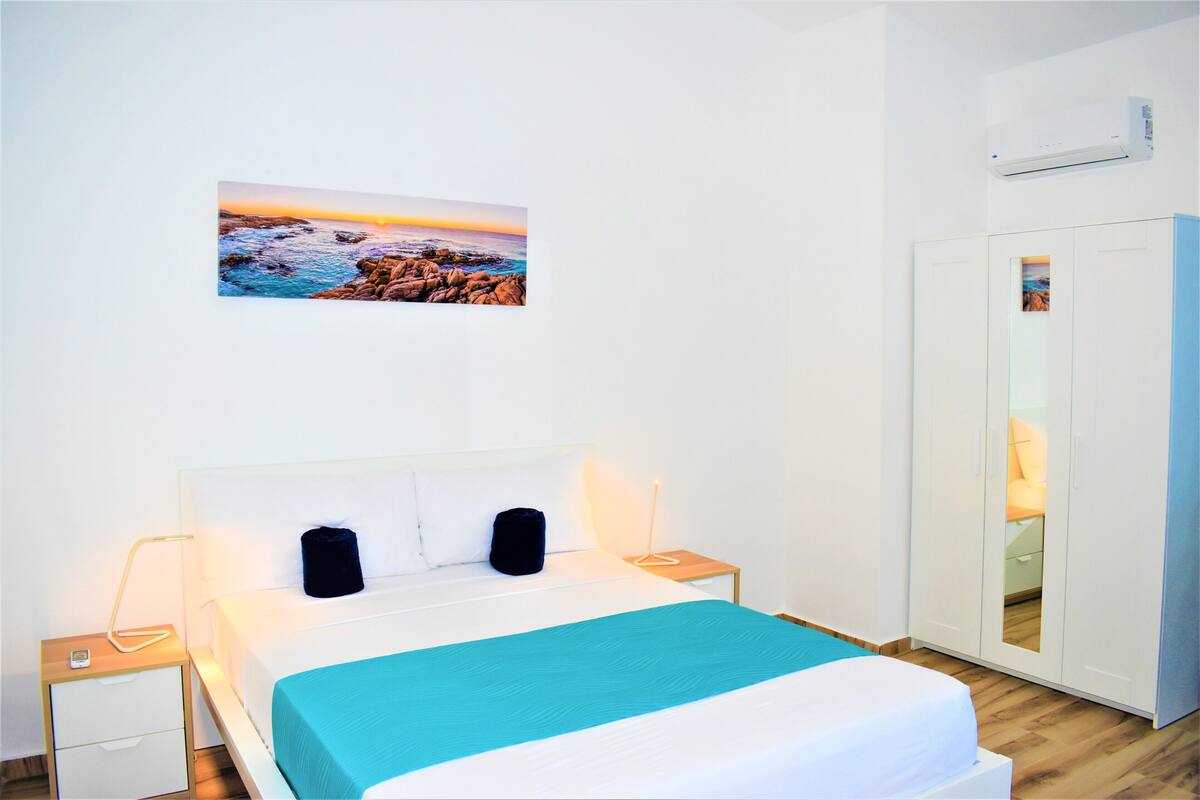A bedroom with a bed, bedside tables, a nightstand, a wardrobe, and a painting of a coastal scene.