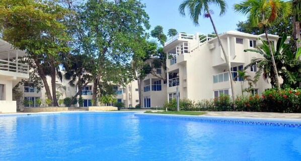 A swimming pool surrounded by multi-story buildings with balconies, trees, and a clear sky. - Tropical Casa Laguna (Cabarete)