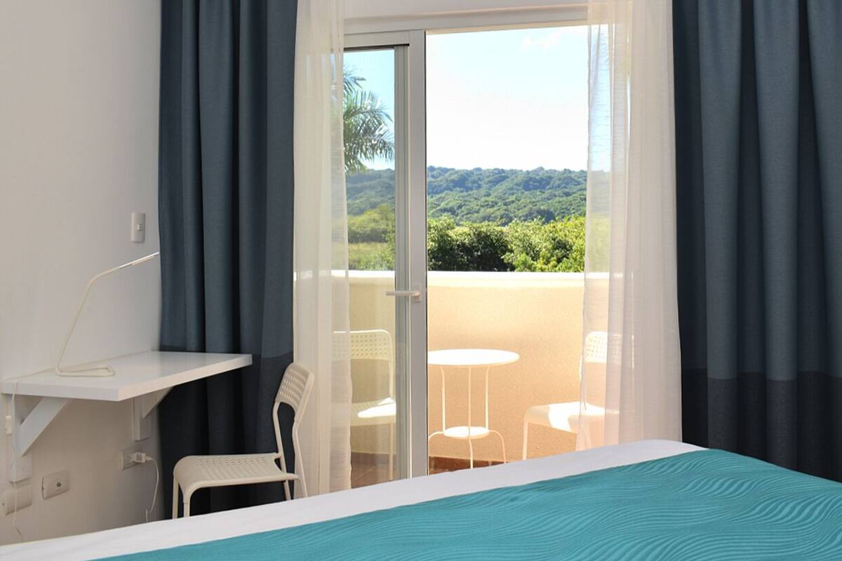 A hotel room with a bed, a desk with a chair, a balcony with a table and chairs, and a view of greenery.