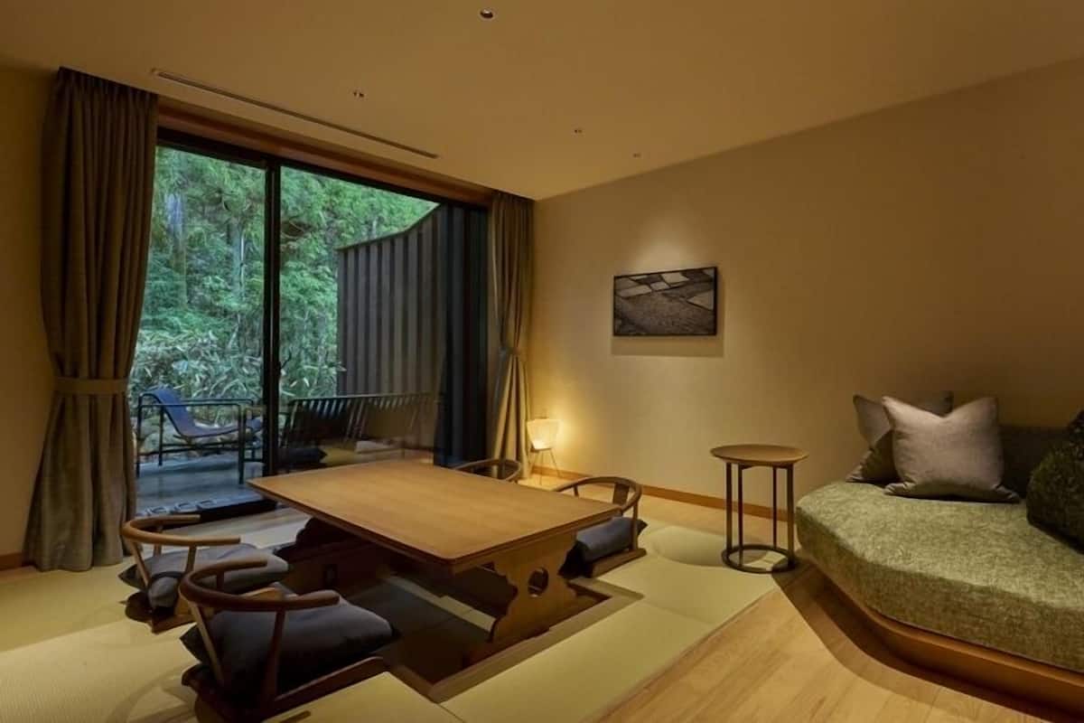 A modern room with a dining table, chairs, a sofa, and a view of greenery through large windows.