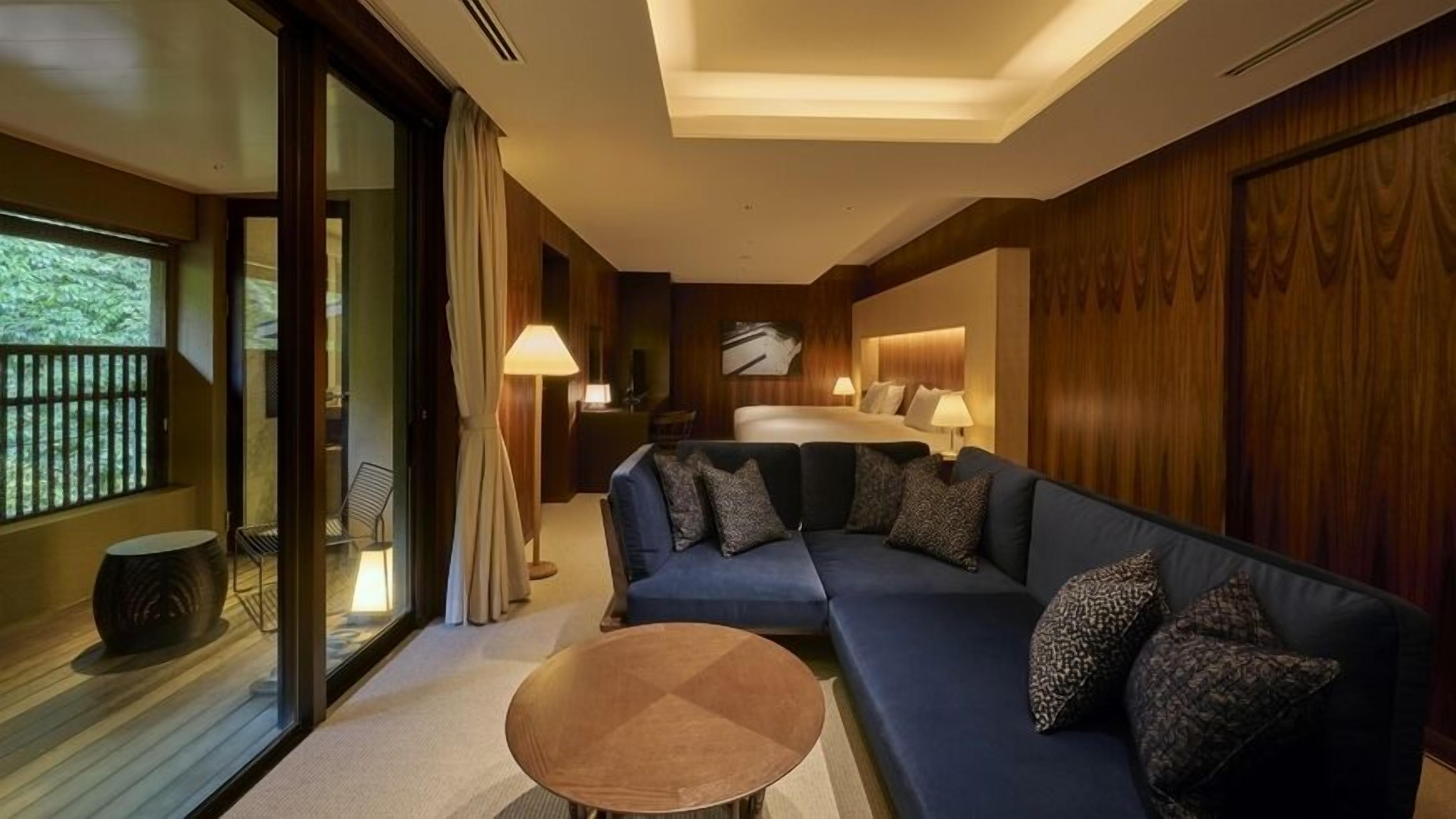 A modern hotel room with a large bed, a sofa, a coffee table, and a view of greenery through the window.