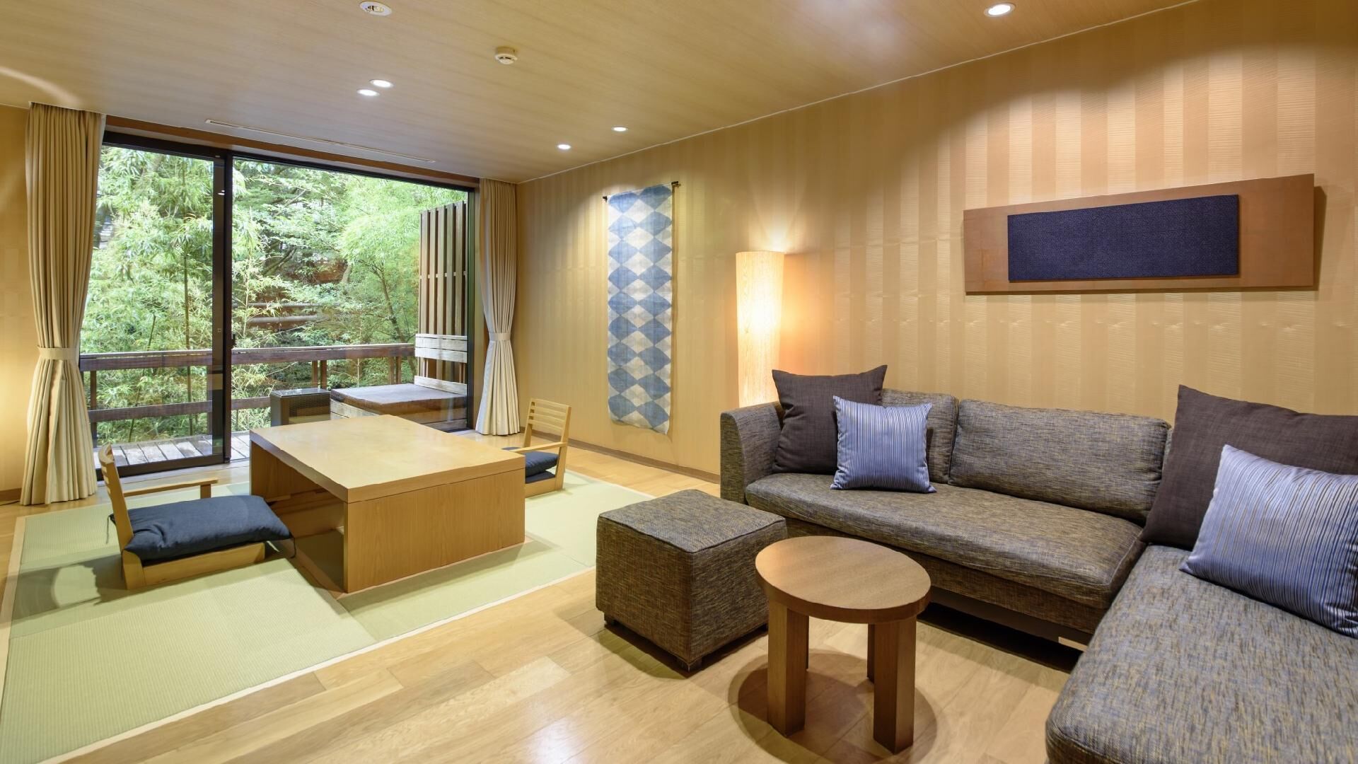 A modern living room with a sofa, ottoman, and a table, large windows with curtains, and a view of greenery.