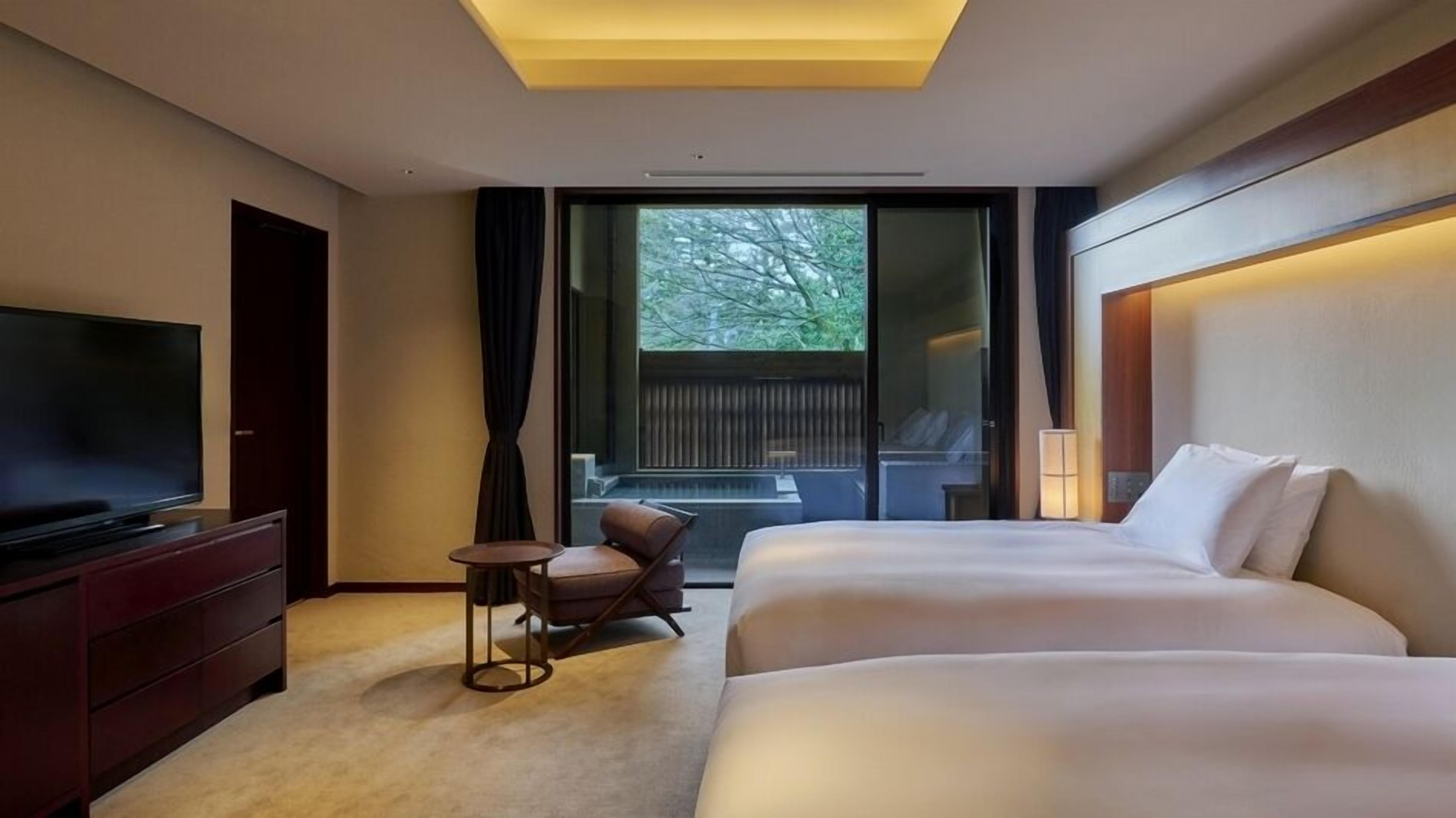 A hotel room with a TV, two beds, a small table, a chair, a window with a view of trees, and a ceiling light.
