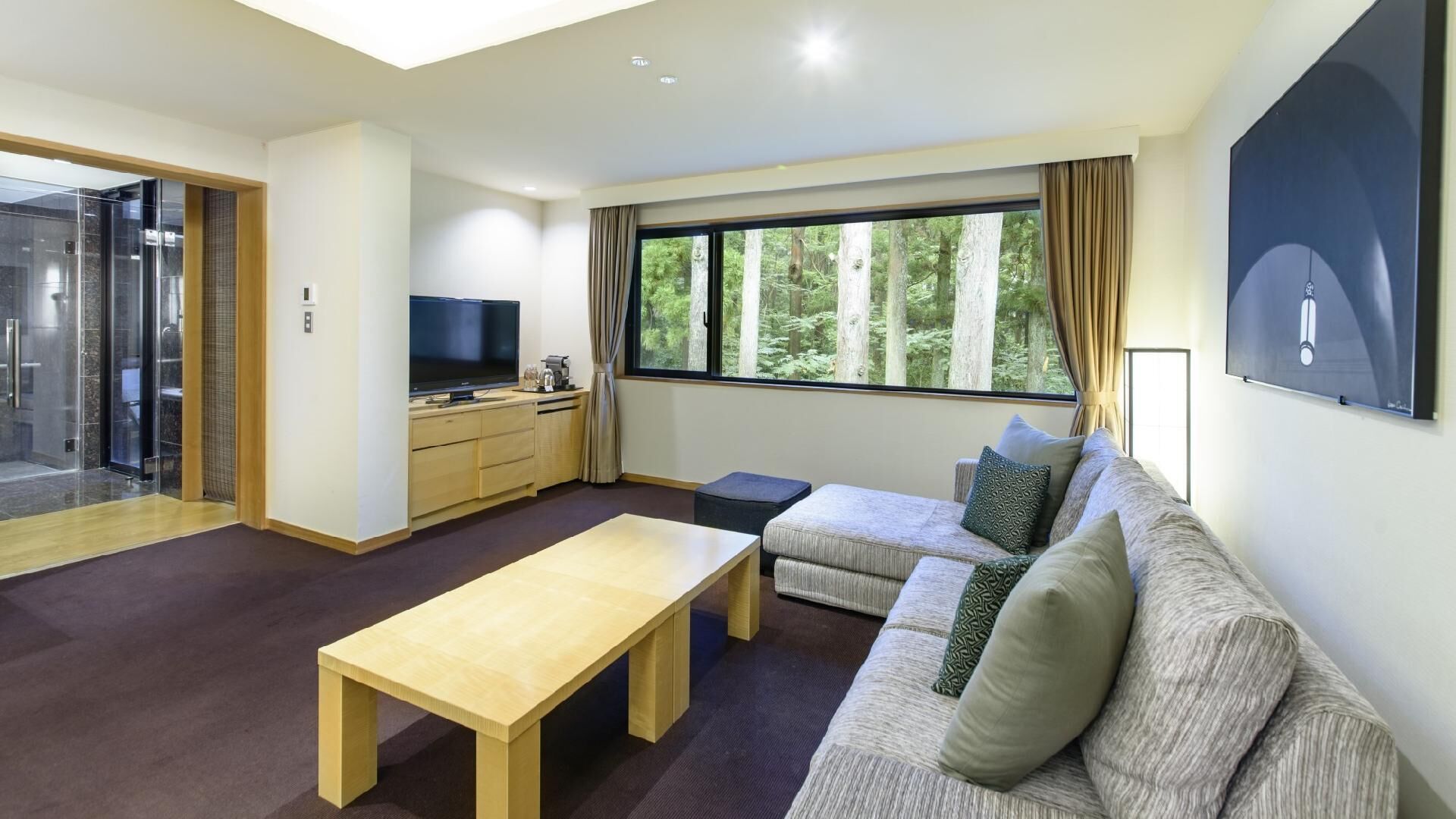A modern living room with a sofa, a coffee table, a TV, and a large window with a forest view.