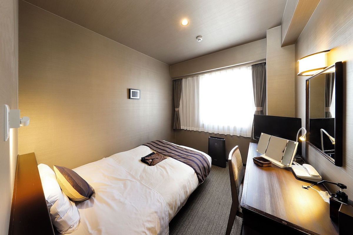 A hotel room with a single bed, a desk with a chair, a mirror, and a window with curtains.