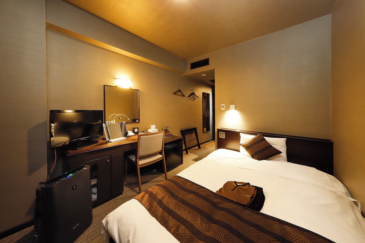 A hotel room with a bed, bedside table, lamp, desk with computer, chair, and a television.