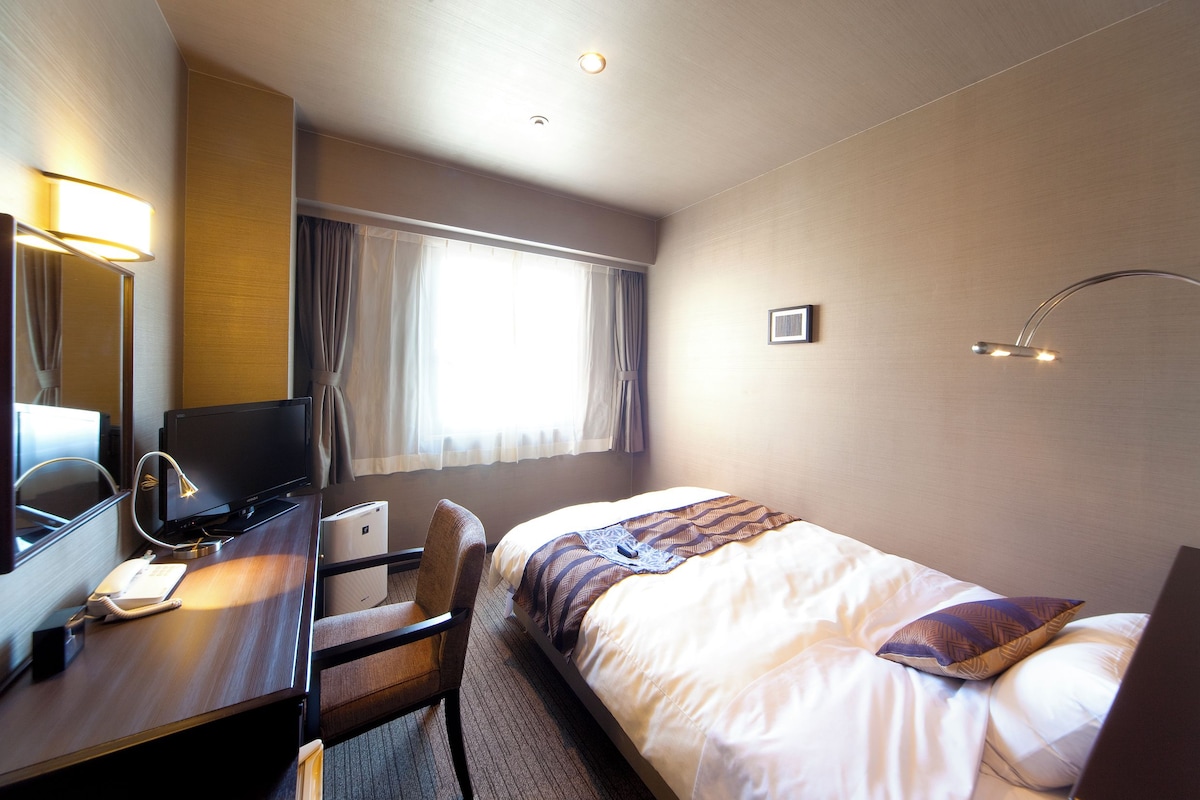 A hotel room with a bed, a desk with a computer, a chair, a desk lamp, a telephone, a window with curtains, and a wall-mounted picture.
