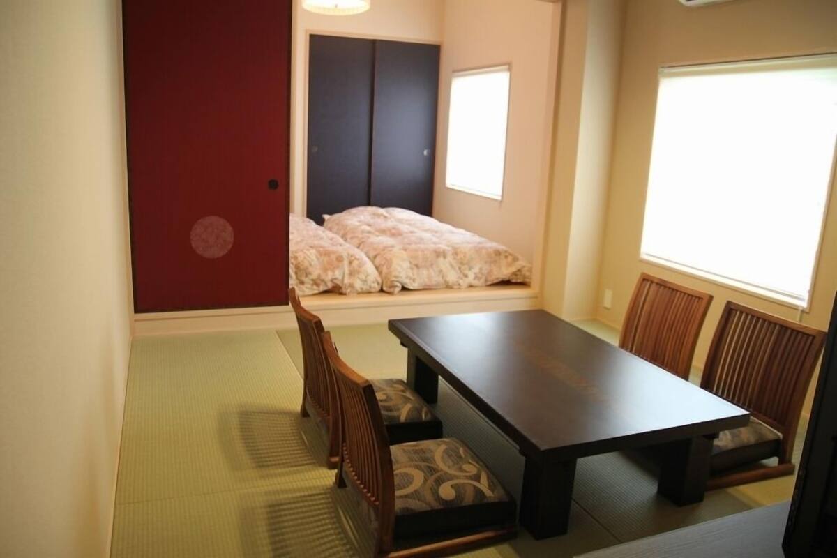 Una habitación con suelo de tatami, una mesa baja, sillas de madera, una puerta corredera y un ventanal.