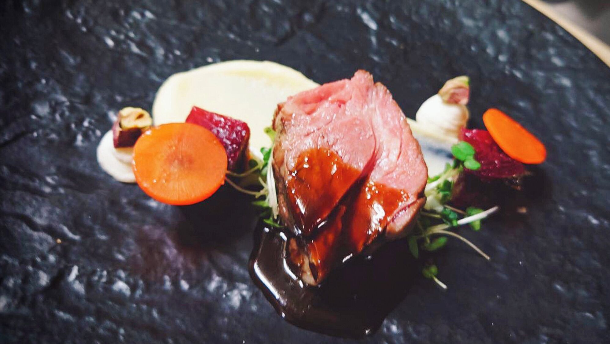 A dish with a slice of meat, a carrot, and some greens on a dark surface.