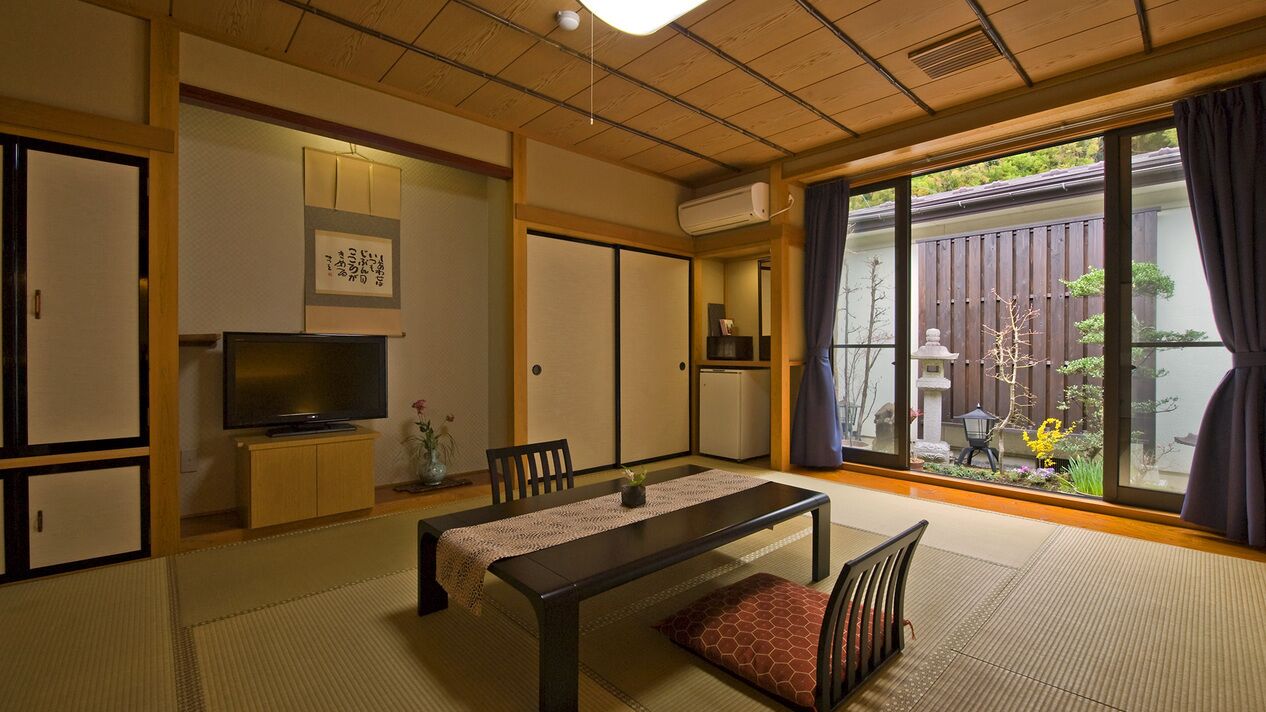 Ein traditioneller japanischer Raum mit Tatami-Boden, einem niedrigen Tisch mit Kissen, einem Fernseher, Schiebetüren und Blick auf einen Garten.
