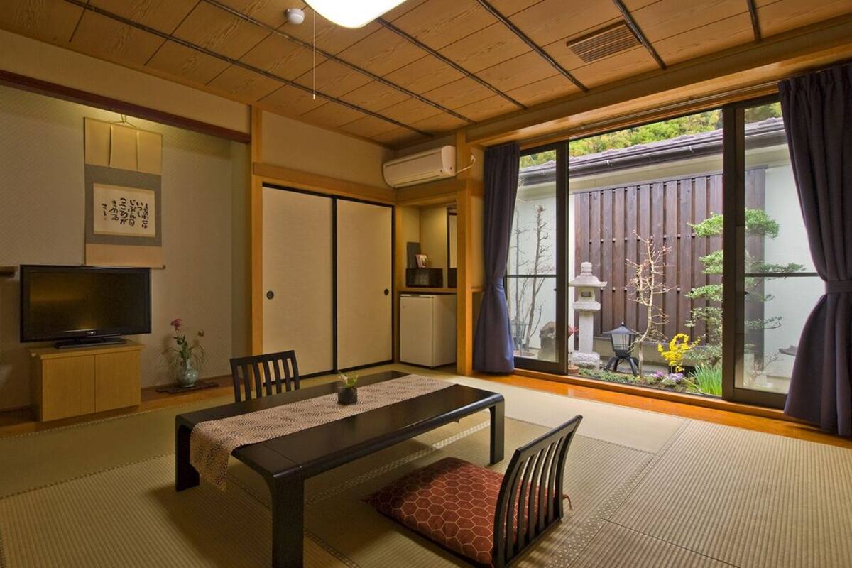 Une pièce de style japonais traditionnel, avec un sol recouvert de tatamis, une petite table basse avec des coussins, un téléviseur, des portes coulissantes et une vue sur un jardin.