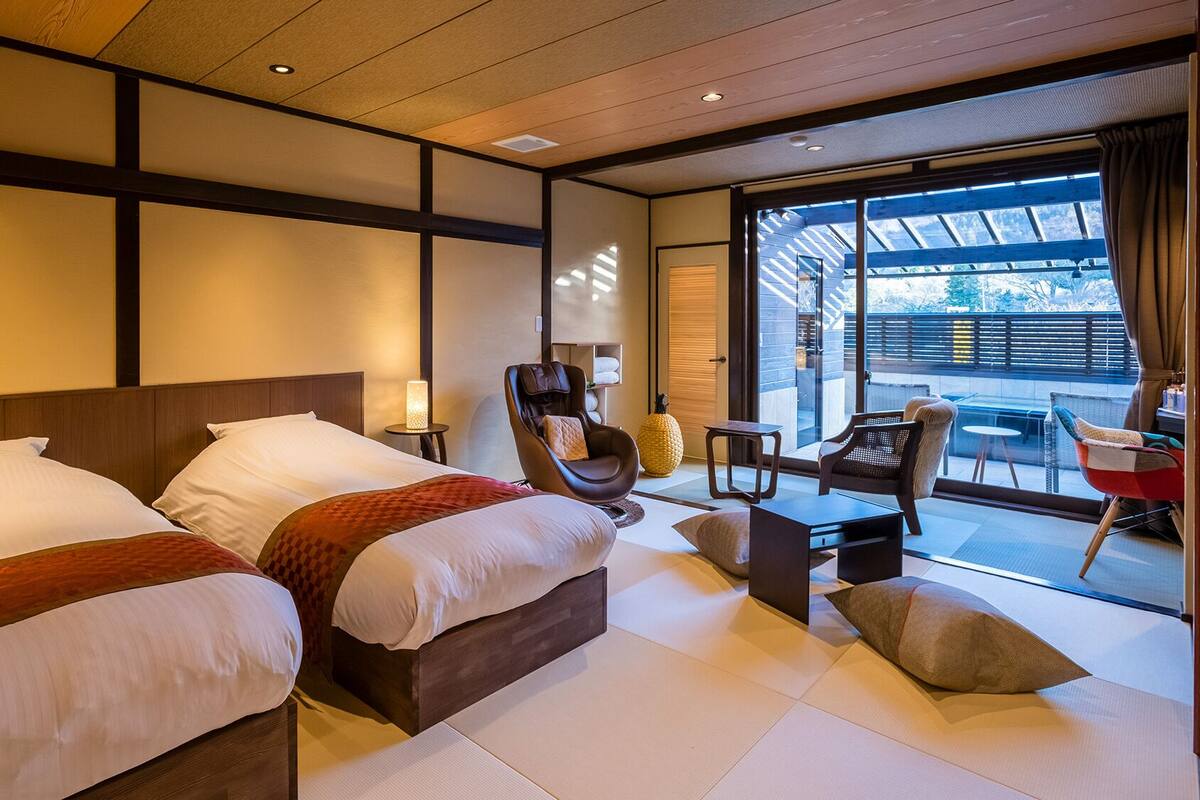 Une chambre d’hôtel avec deux lits, un fauteuil massant, une petite table et un balcon avec des sièges.