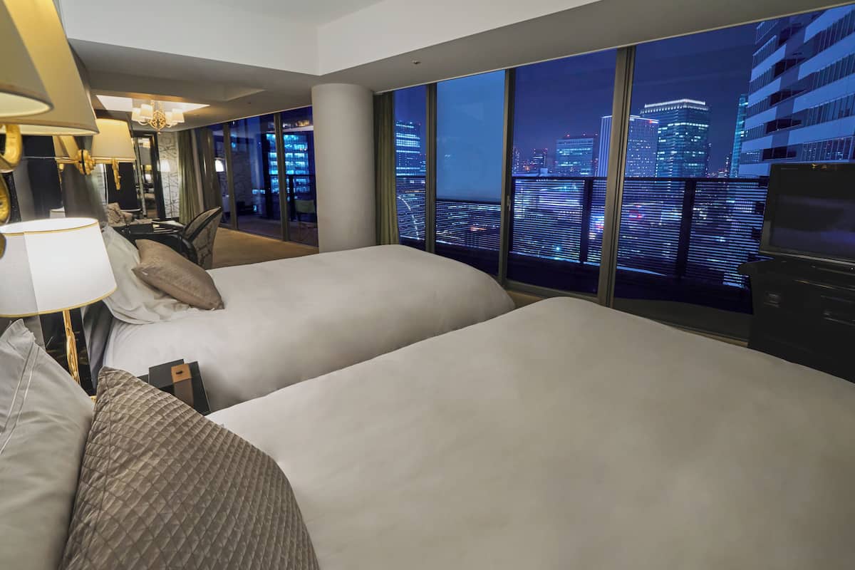 A hotel room with two beds, a large window with a city view, a desk with a chair, and a television.