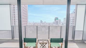 A balcony with two green chairs and a small table, overlooking a cityscape with high-rise buildings.