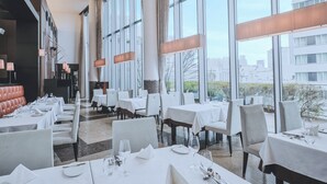 Moderni ravintola, jossa on suuret ikkunat, valkoiset pöydät ja nahkaverhoiltu istuma-alue.