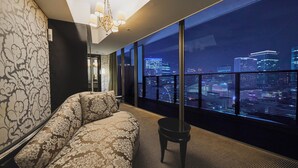 Moderni hotellihuone, jossa on suuri sänky, pieni pöytä ja näkymä kaupunkiin suurista, lattiasta kattoon ulottuvista ikkunoista.