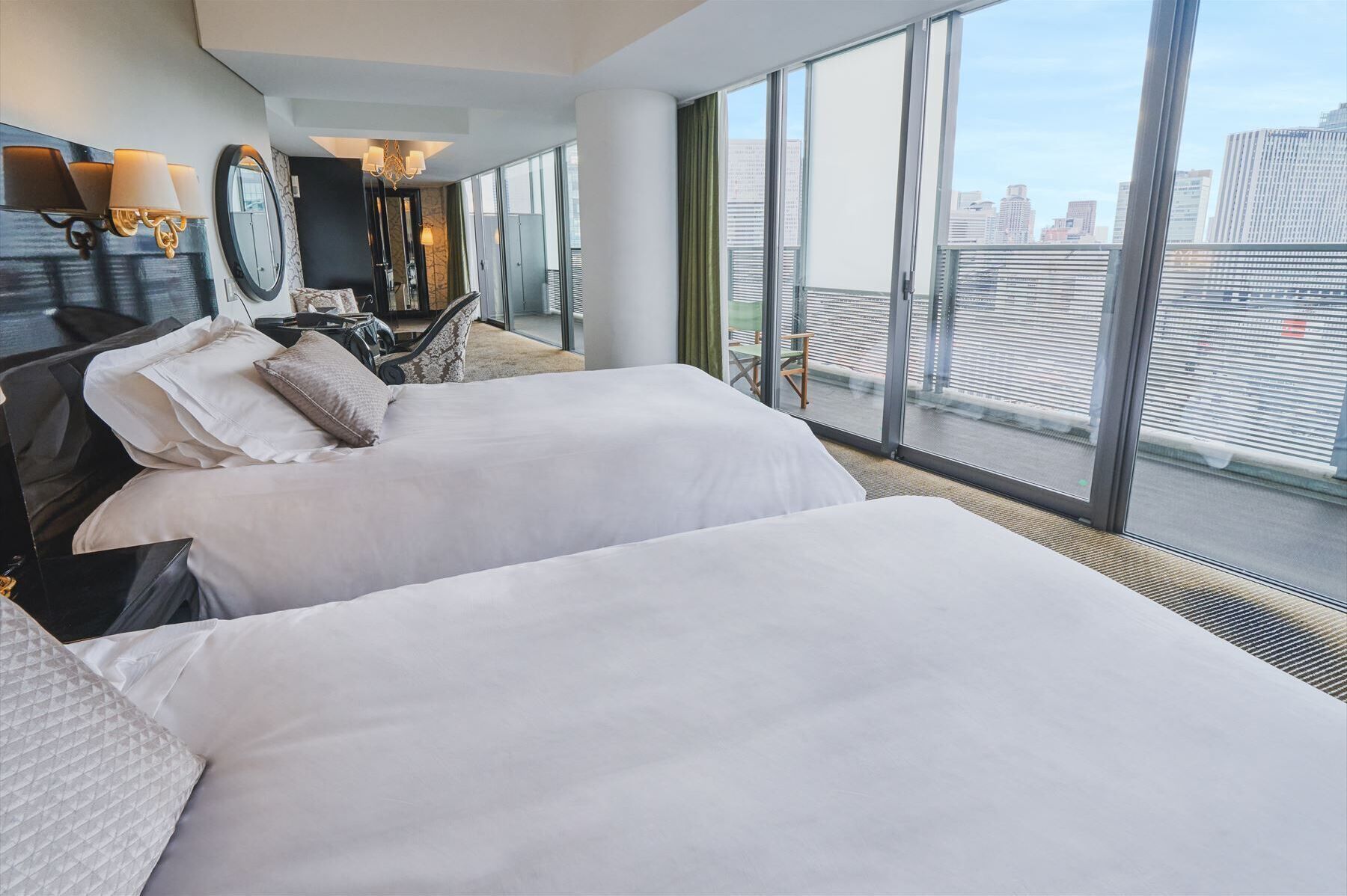 A hotel room with two beds, a large window with a city view, a desk with a chair, and a mirror.