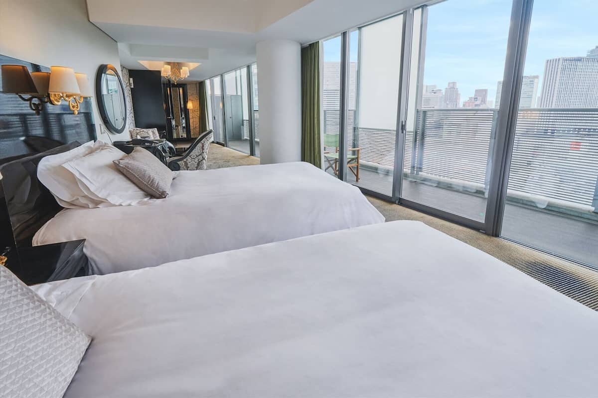 A hotel room with two beds, a large window with a city view, a desk with a chair, and a mirror.