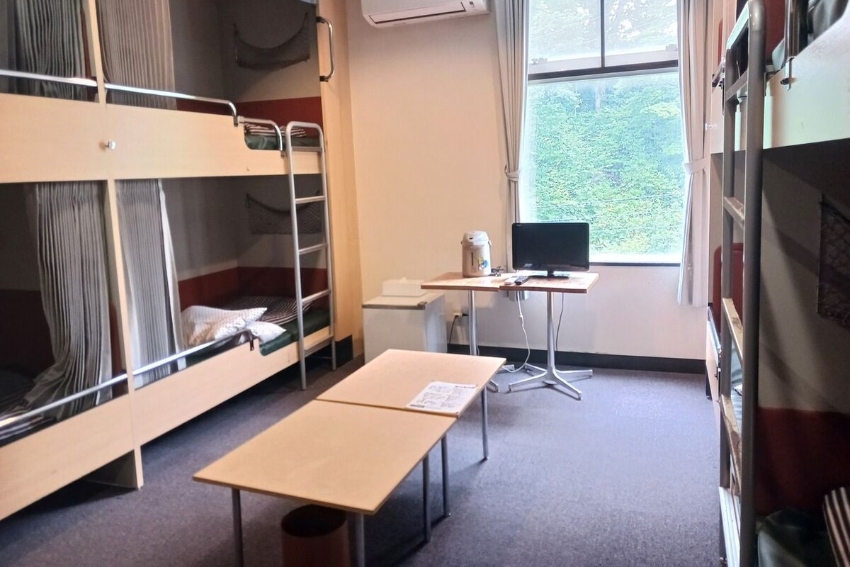 Een studentenkamer met stapelbedden, een bureau, een computer en een raam met uitzicht op groen.