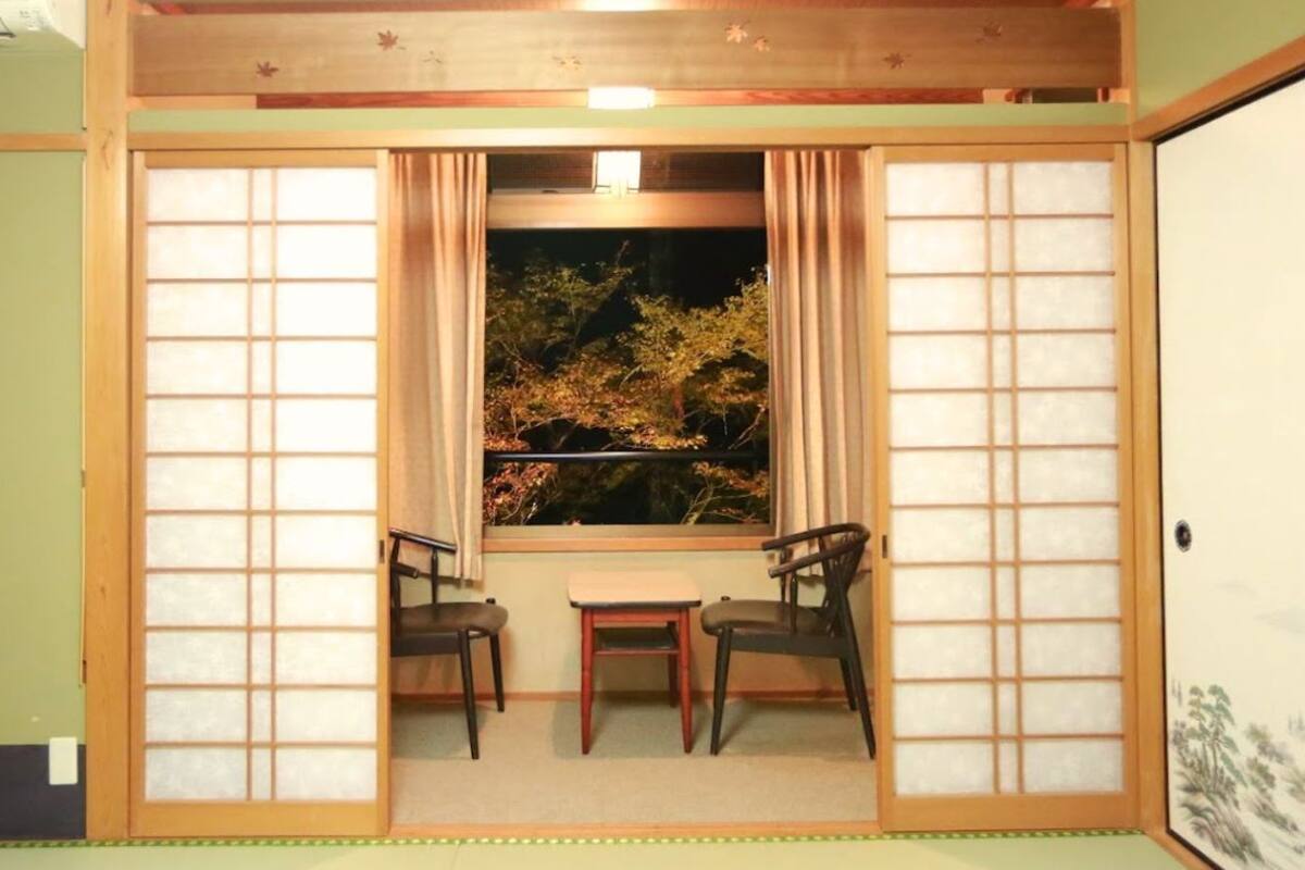 Una habitación japonesa tradicional con suelo de tatami, una mesita baja, dos sillas y una ventana con vistas a árboles otoñales.
