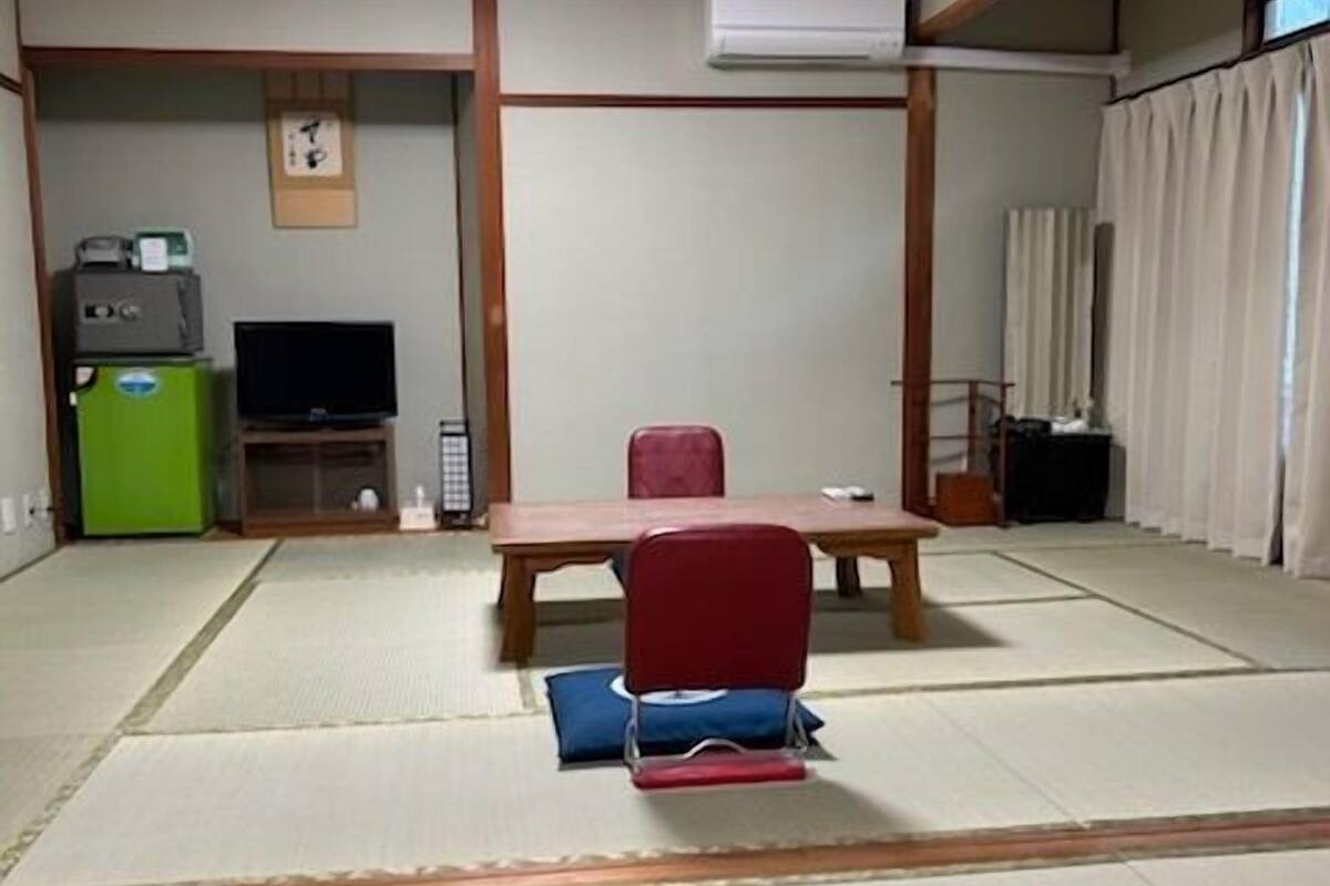 Una habitación con suelo de tatami, televisión, un refrigerador pequeño, una silla roja y una mesa de madera.
