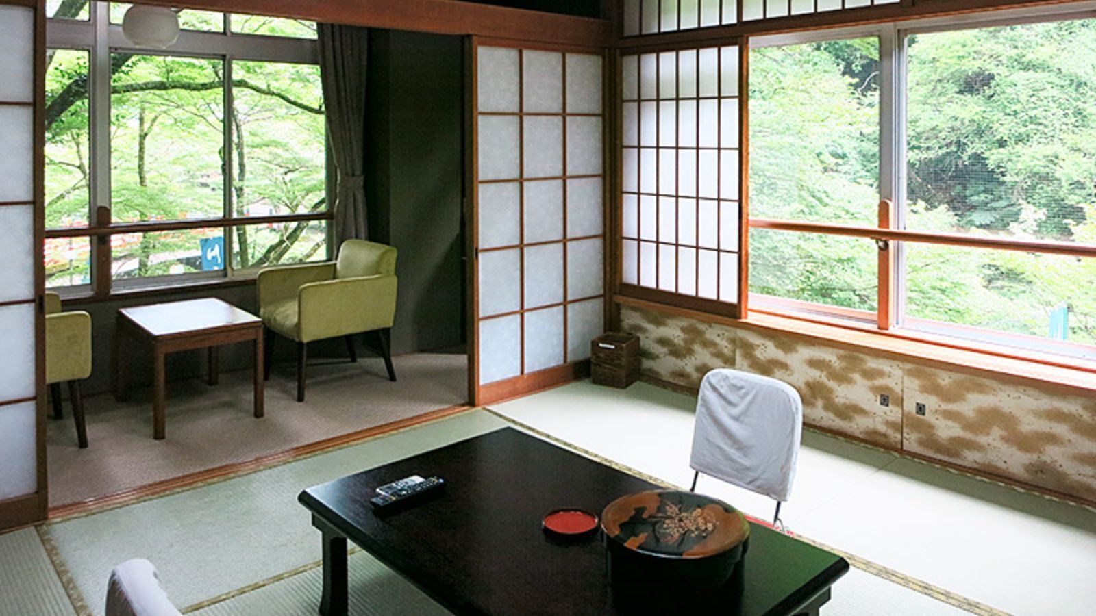 Um quarto japonês tradicional com tatames, uma mesa baixa com controle remoto, uma cadeira e vista para o verde lá fora.