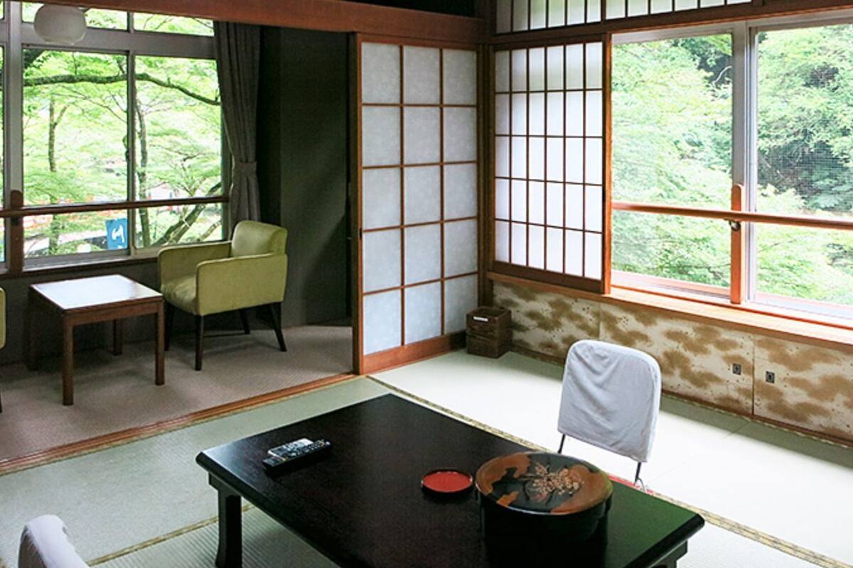 Una habitación tradicional japonesa con suelo de tatami, una mesa baja con un control remoto, una silla y vistas al exterior con vegetación.