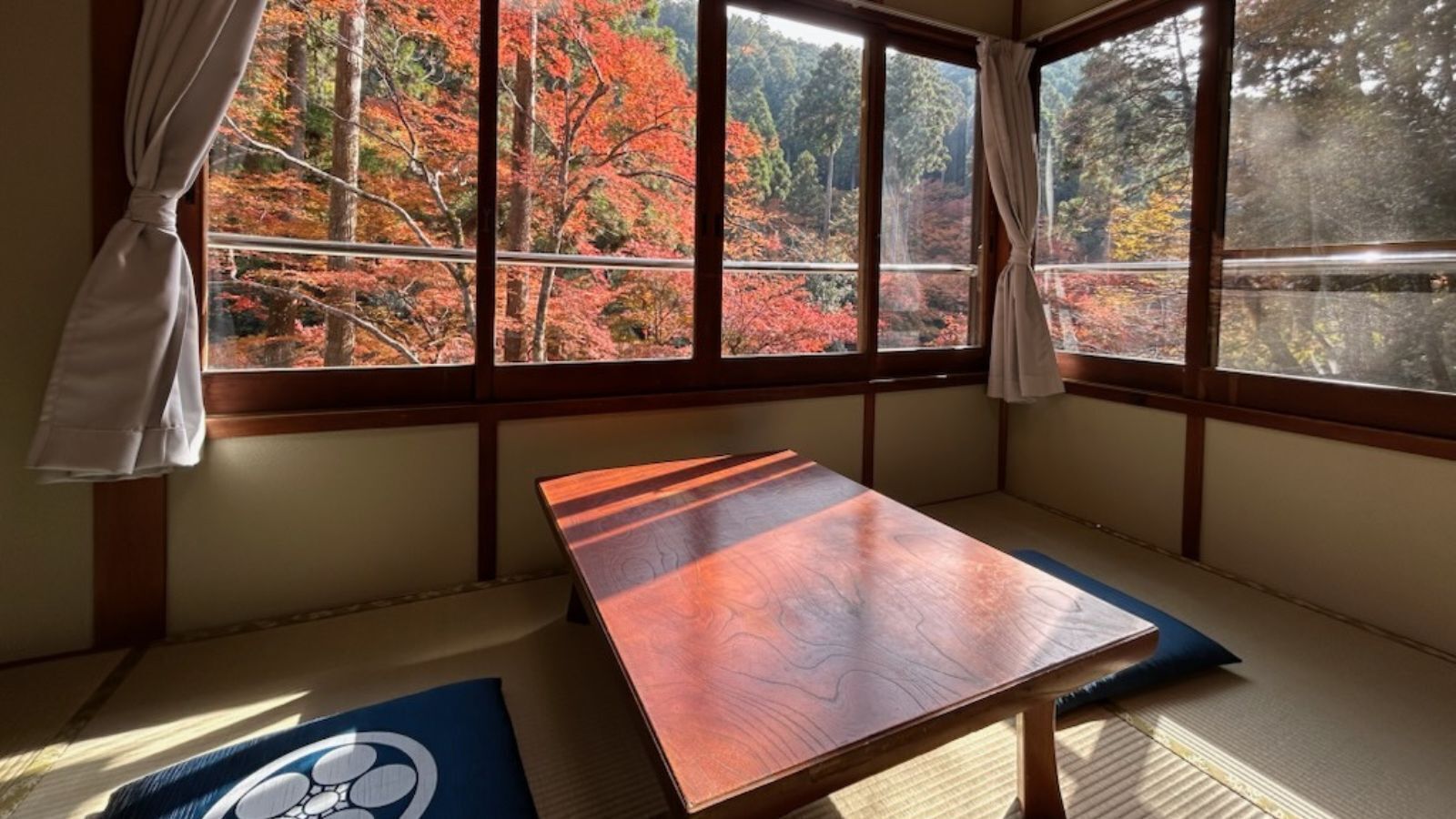 Quarto com tatames, uma mesa de madeira baixa, tatames e vista para árvores de outono através de grandes janelas.