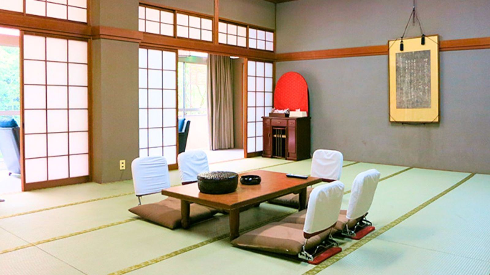 Um espaço com tatames, uma mesa baixa e cadeiras, um nicho vermelho e um quadro na parede.