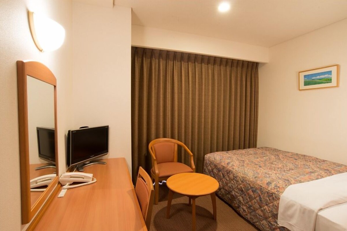 A hotel room with a bed, a desk with a television, a chair, and a window with curtains.