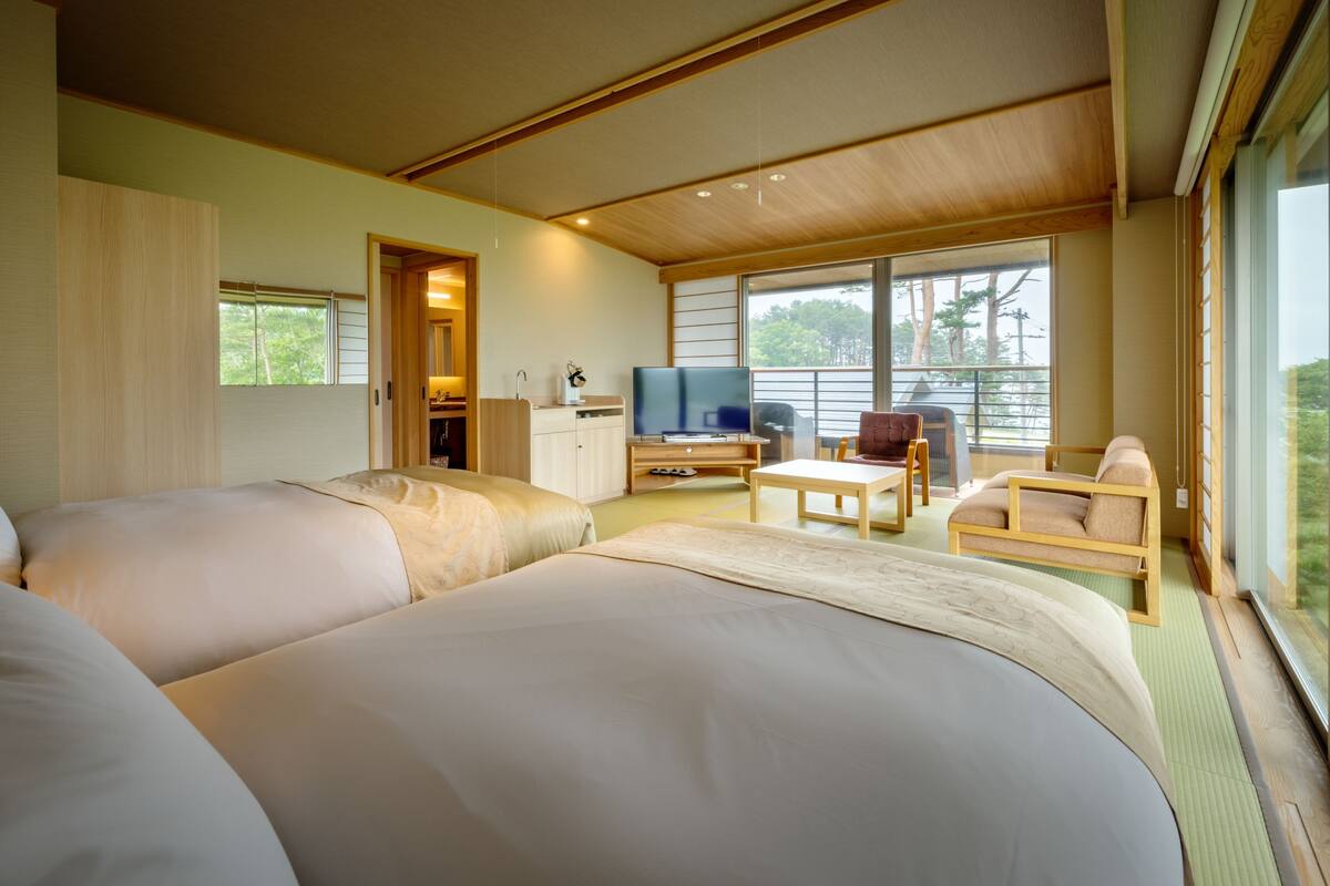 A hotel room with two beds, a desk, a TV, and a balcony with a view of trees.