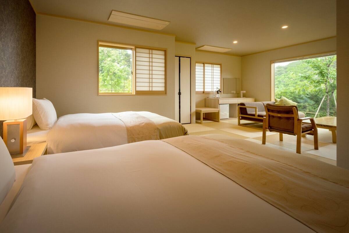 A hotel room with two beds, a desk, a chair, and a view of greenery.