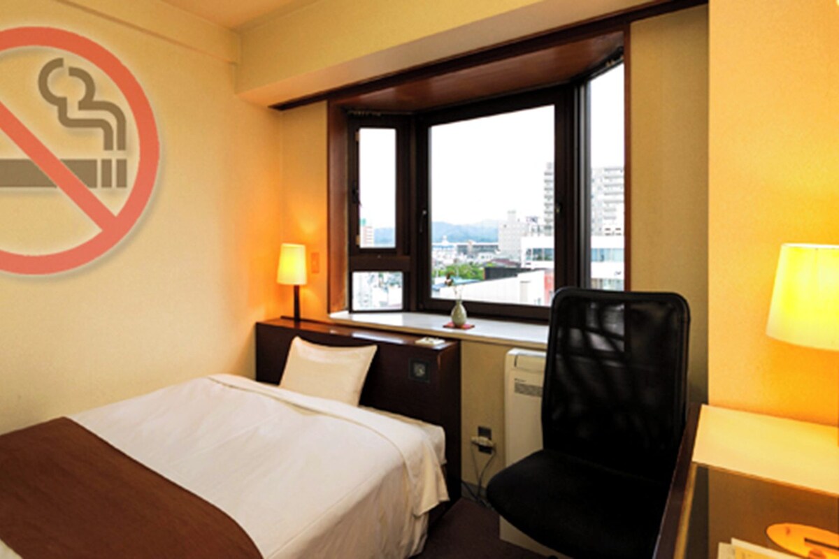 A hotel room with a bed, a desk, a chair, a lamp, and a window with a view of buildings.