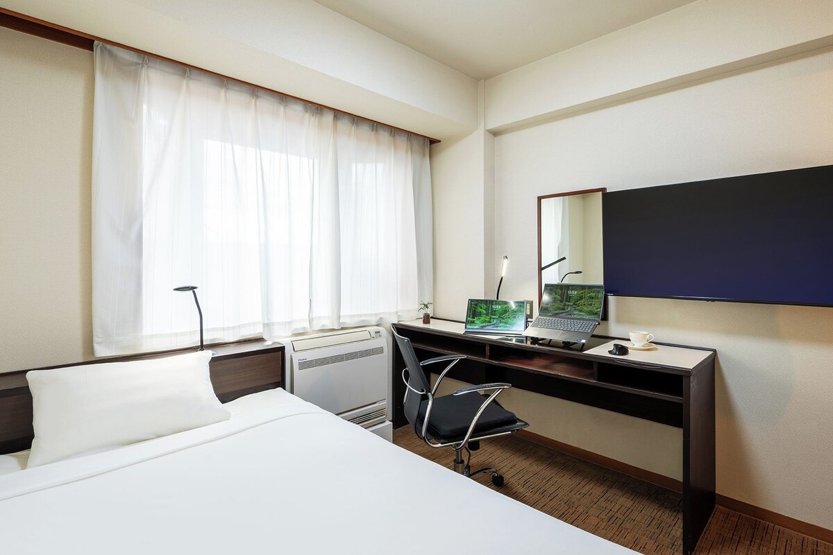 A hotel room with a bed, a desk with a laptop, a chair, a desk lamp, and a large mirror.