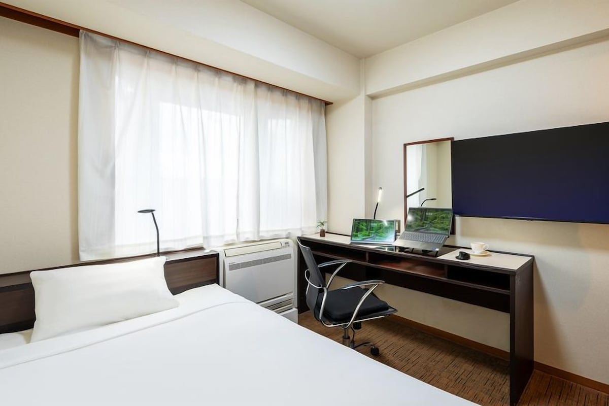 A hotel room with a bed, a desk with a laptop, a chair, a large mirror, and a window with curtains.