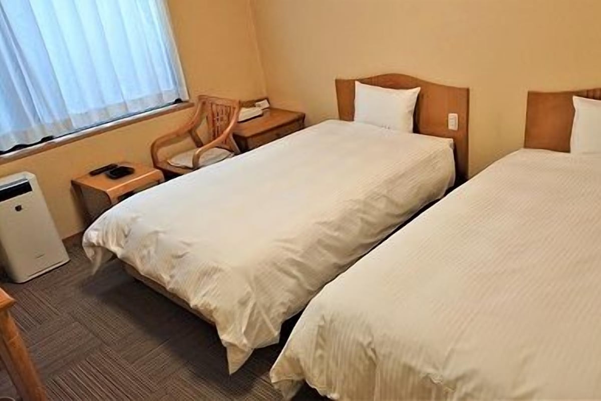 A hotel room with two beds, a desk, a chair, and a small heater.