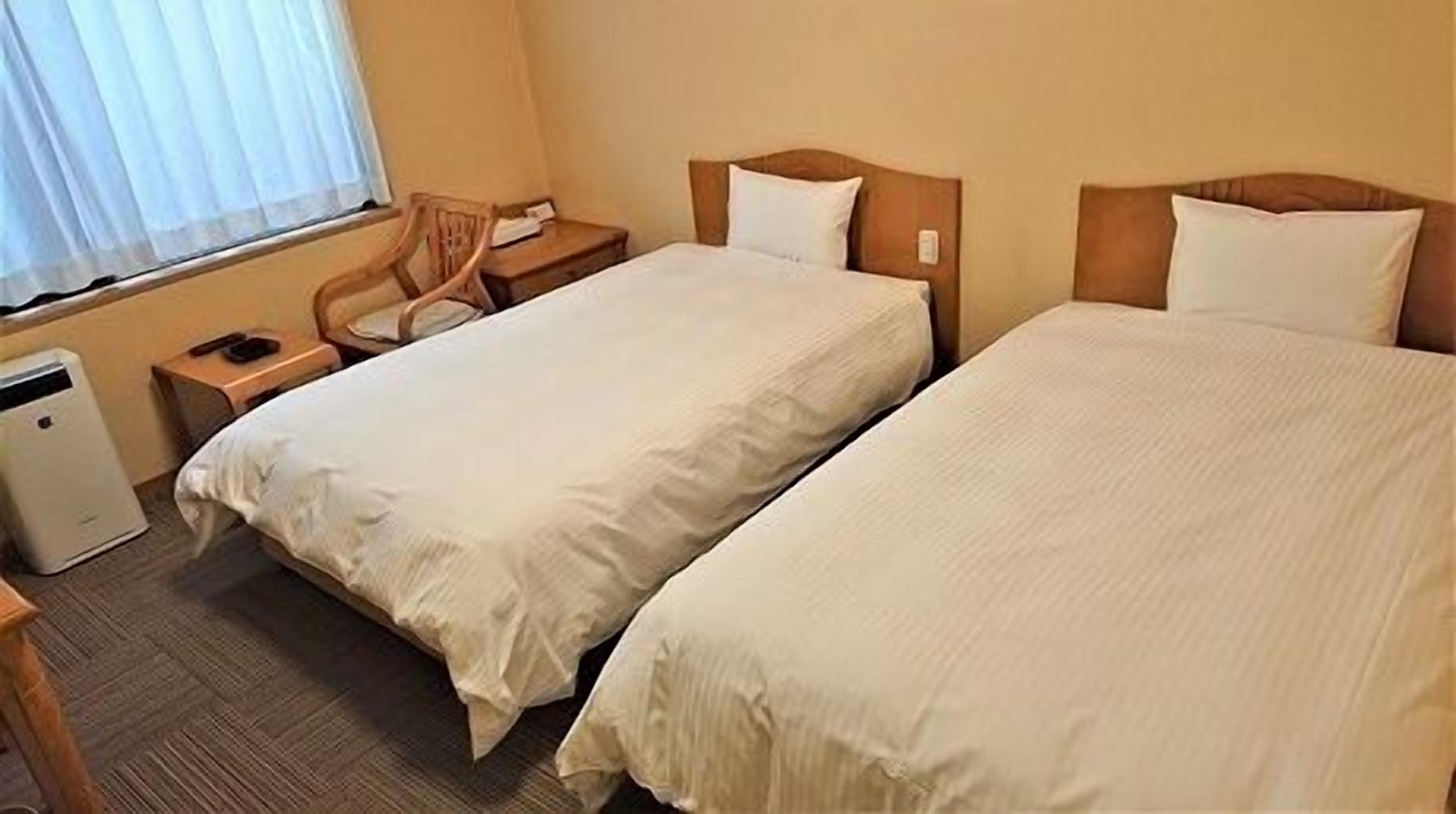 A hotel room with two beds, a desk, a chair, and a small heater.