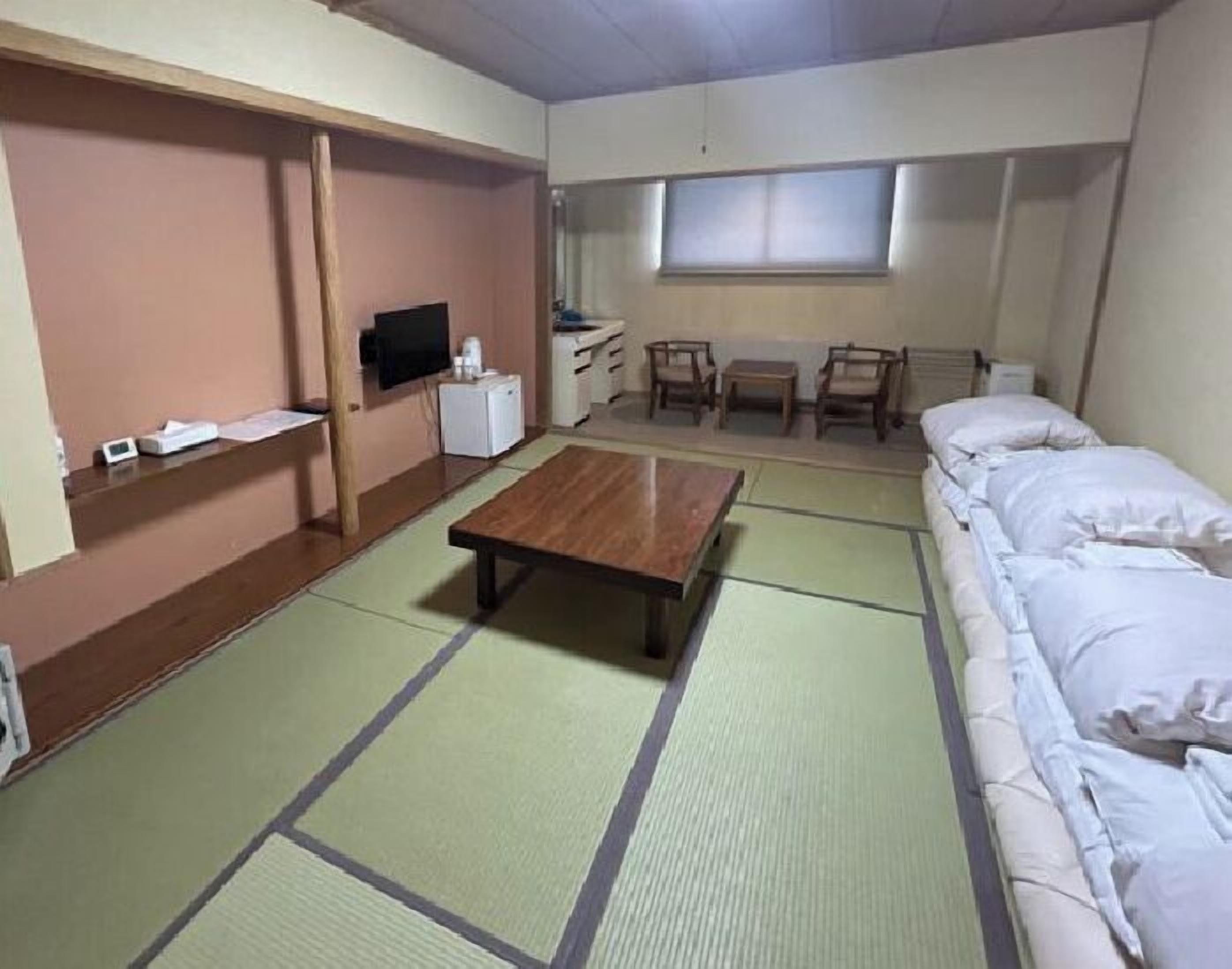 A room with tatami mats, a bed, a TV, a small table, and a desk with chairs.