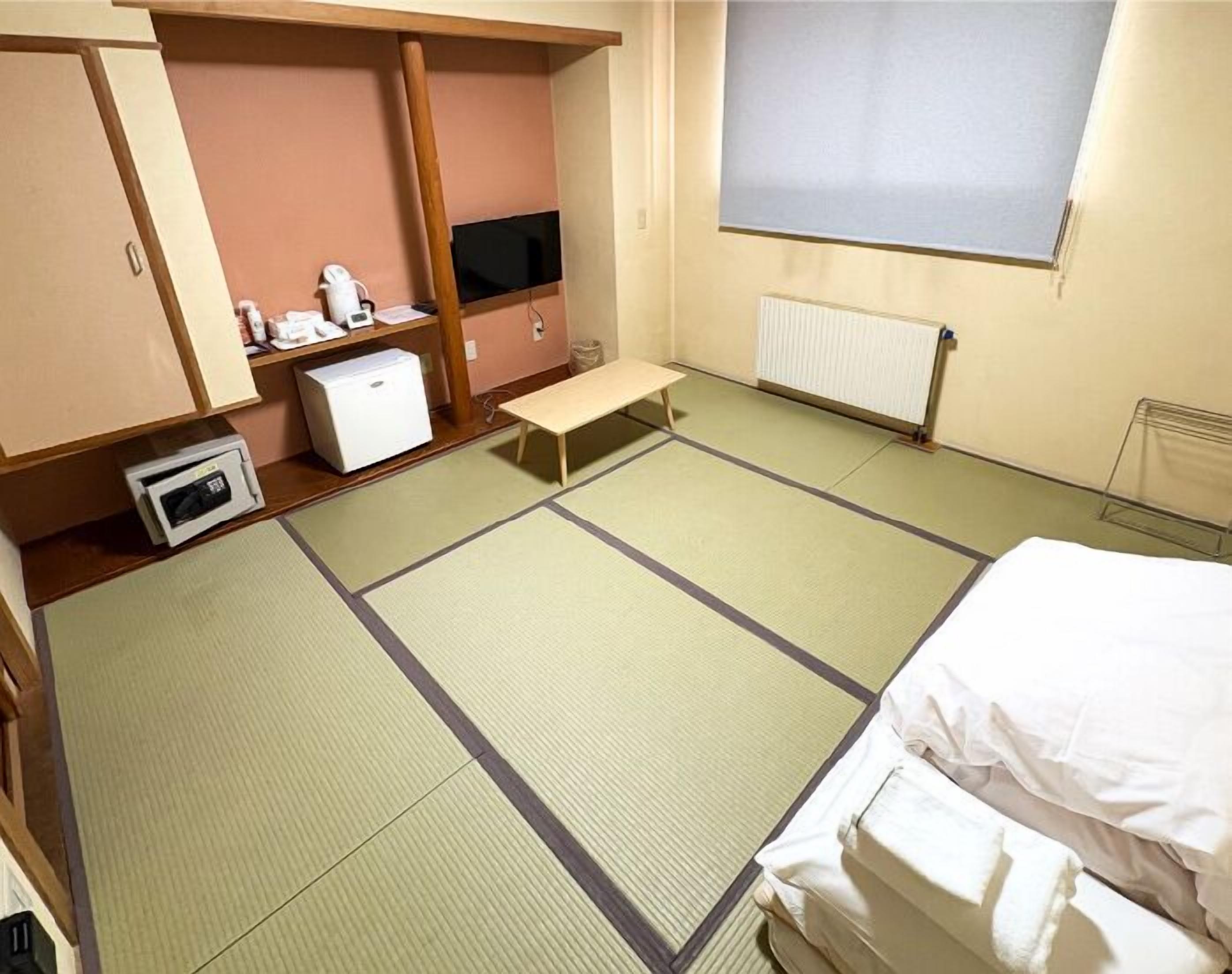 Habitació d'hotel amb sòl de tatami, un llit, un escriptori petit amb un ordinador, una televisió i un radiador.
