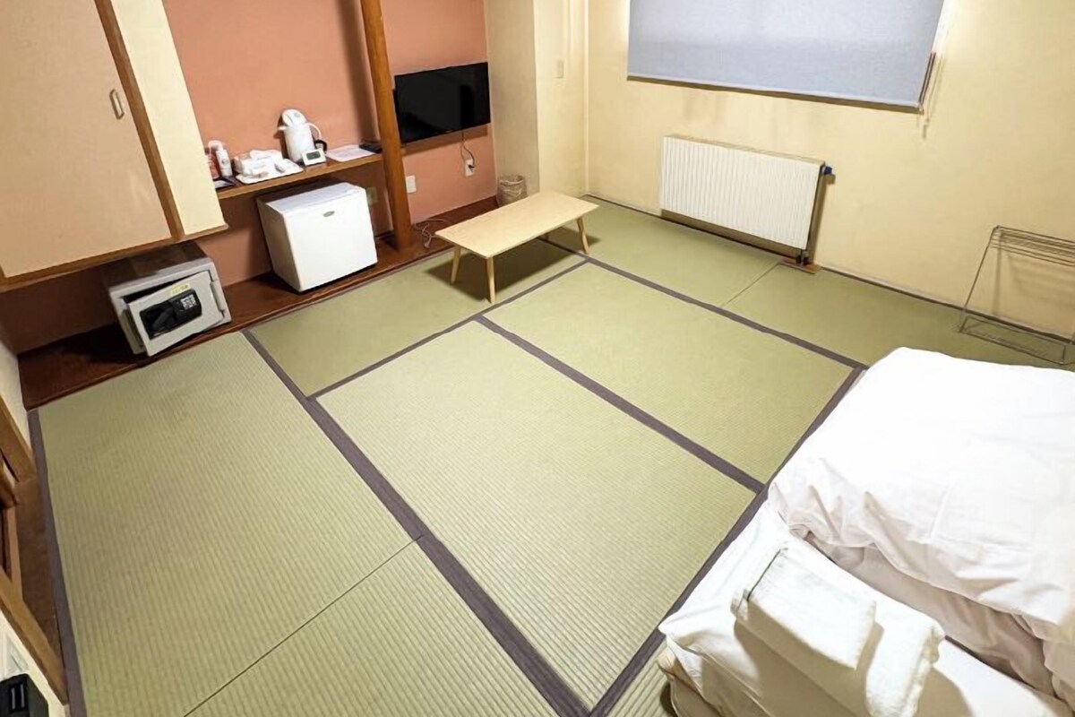 A room with tatami flooring, a bed, a small desk with a computer, a television, and a radiator.
