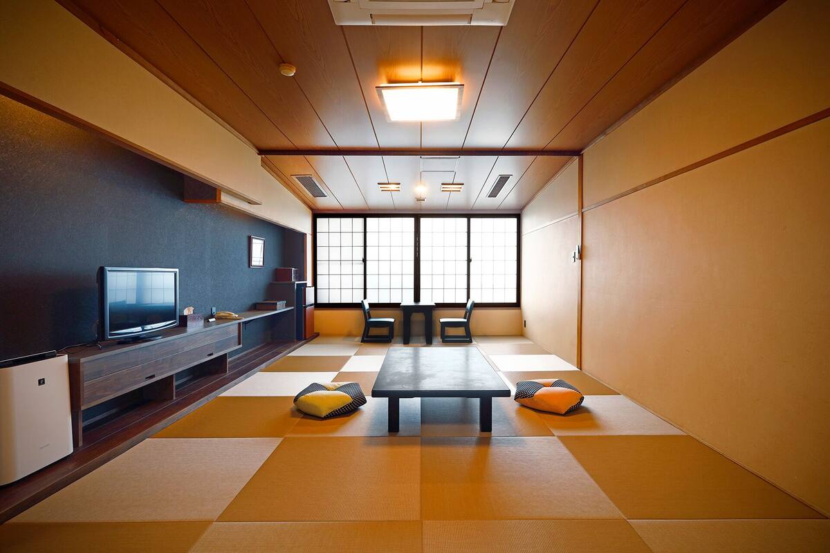Une pièce avec des tatamis, une télévision, une table, des chaises et une grande fenêtre.