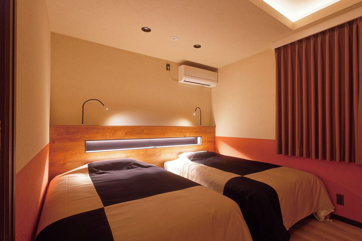 Habitación de hotel con dos camas, una lámpara en el techo, un equipo de aire acondicionado y una ventana con cortinas.