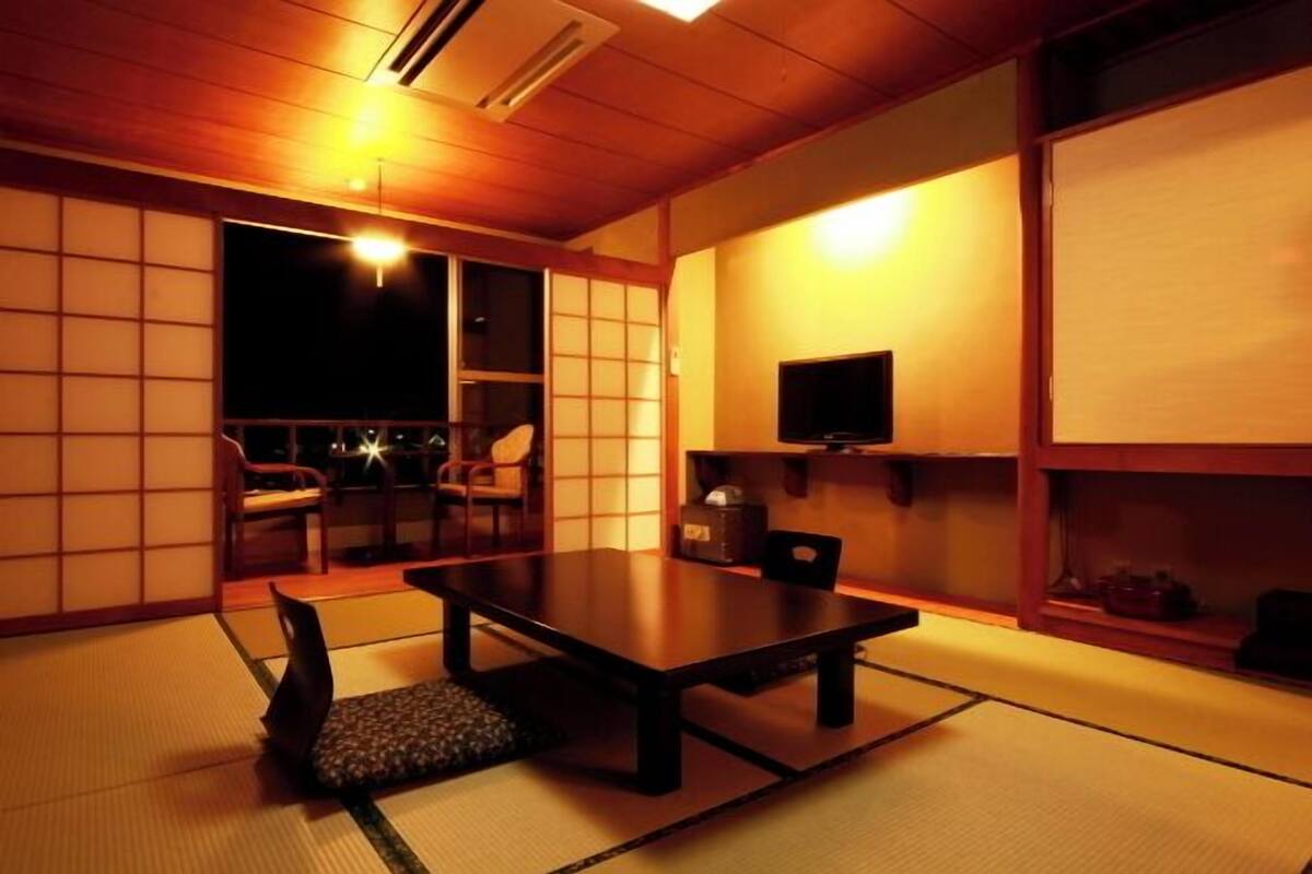 Une pièce de style japonais traditionnel, avec un sol recouvert de tatamis, une table basse, des chaises, un téléviseur et des portes coulissantes.